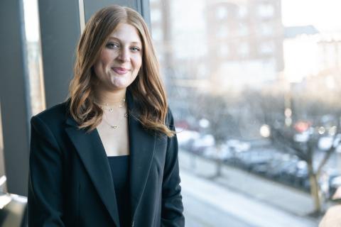 Julia Klayman poses next to a window for a portrait wearing a black suit jacket and a demure smile.